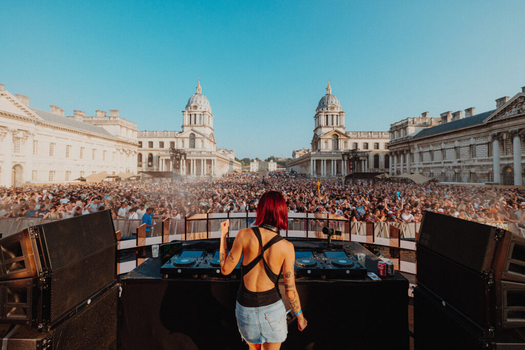 A Panoramic Shot the outside of the Old Royal Naval College with a red headed DJ behind DJ decks and a huge crowd of people all in the centre of the Old Royal Naval College Grounds